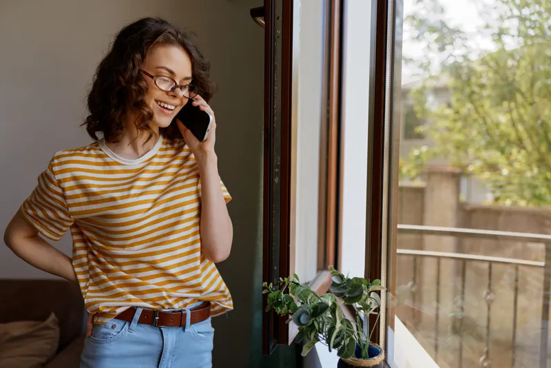 Smiling woman with striped shirt talking on phone by a sunlit window, indoor plant on the sill, casual and cozy home setting, warm vibe, everyday communication.
