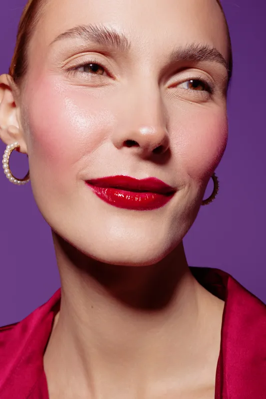 Close-up portrait of a smiling woman with red lipstick and pearl earrings on purple background. Concept of beauty, makeup, fashion, and confident female model with smooth skin and elegance.