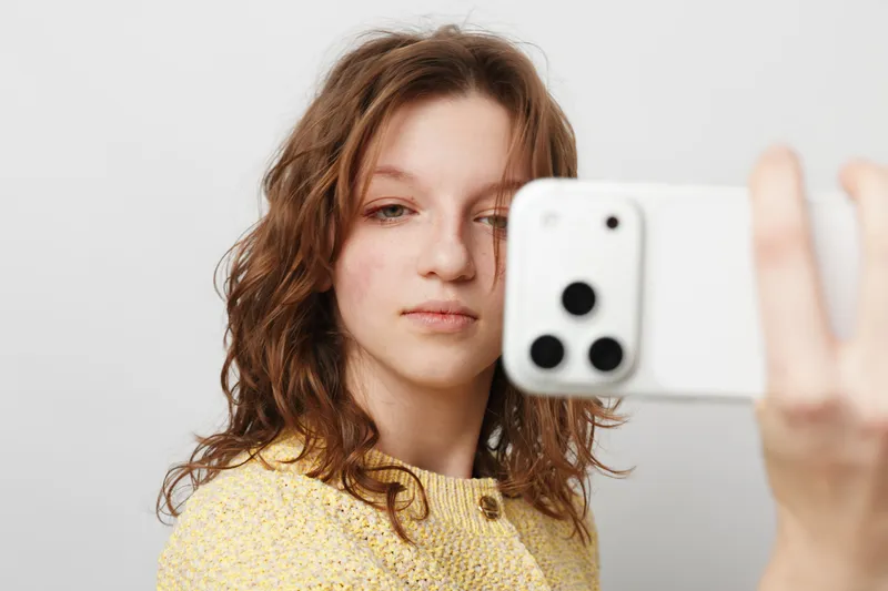 Young woman with curly hair takes a selfie with a smartphone on white background. Casual style, natural expression, and soft lighting create a relaxed mood.