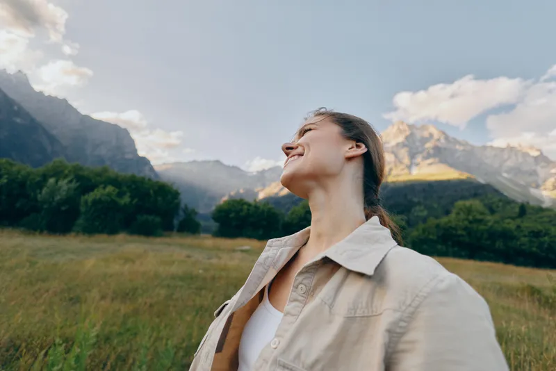 Woman enjoying outdoor life in a wide meadow with distant mountains, wearing light jacket, looking up with a joyful smile, embodying relaxed natural lifestyle.