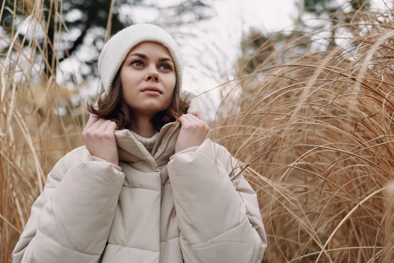 Woman in a warm coat and beanie among tall grasses, outdoor winter scene with soft natural light and calm, cozy mood.