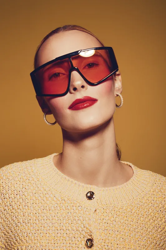 Stylish woman with large red sunglasses and red lipstick poses confidently against a warm brown background. She wears gold hoop earrings and a textured beige sweater.