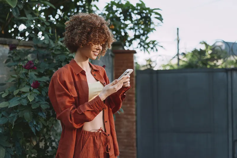 Woman with curly hair in orange casual outfit outdoor, looking at smartphone with a relaxed joyful mood in a garden scene, lifestyle moment, natural light