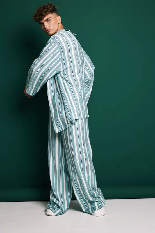 Young man posing in striped loose fit shirt and wide pants with white sneakers against dark green background in studio. Fashion model with curly hair and casual outfit.