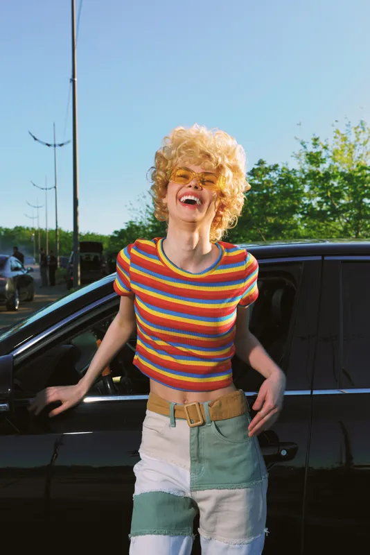 Smiling woman with curly blonde hair wearing a striped tee poses beside a car outdoors. Retro film color vibe, casual fashion, bright mood, carefree energy.