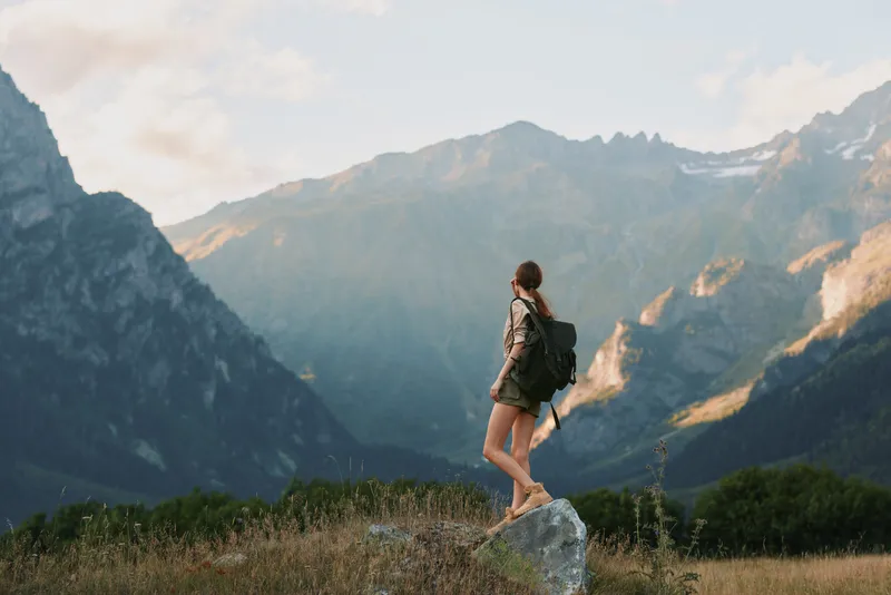 Hiking woman with backpack stands on a rock in a mountain landscape, enjoying nature and solitude, outdoor adventure mood, bright sky. warm sunlight, distant peaks, peaceful