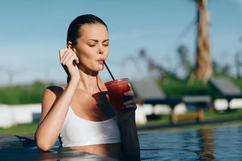 A woman relaxes in a pool, sipping a red beverage through a straw, enjoying sunny outdoor surroundings. A lifestyle moment of refreshment and leisure.
