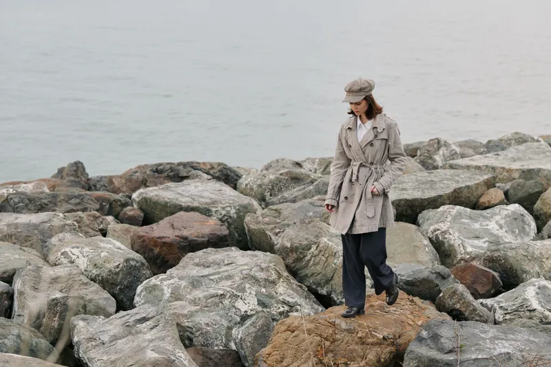 Woman in beige coat and hat walks carefully on large rocks near calm sea. Overcast weather creates a muted atmosphere. Concept of exploration, nature, and solitude by the water.