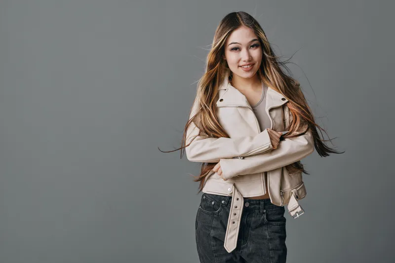 Young woman posing on gray background in trendy light jacket and black jeans, smiling and looking confident with long hair and crossed arms in casual style.