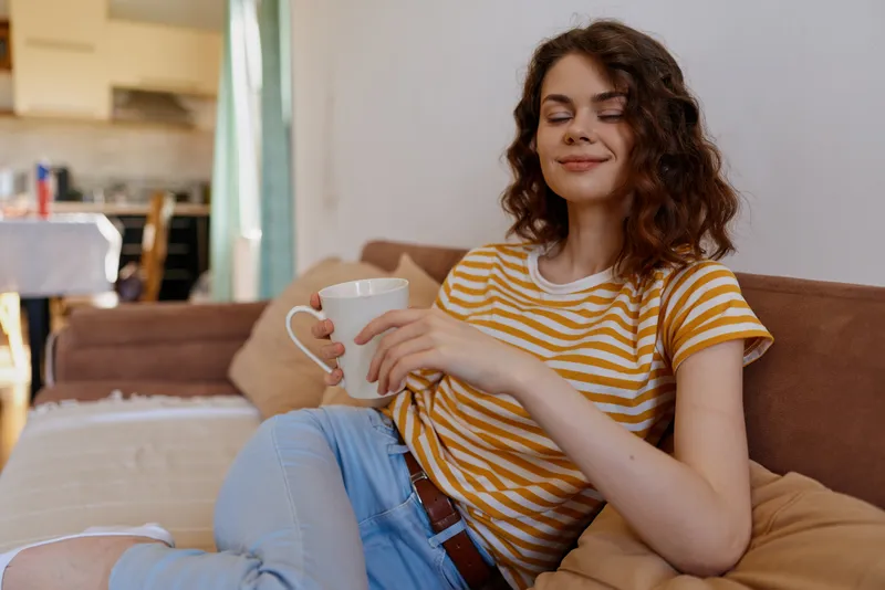 Young woman wearing a striped tee relaxes on a couch at home, holding a mug and smiling softly in a cozy living room with natural light.