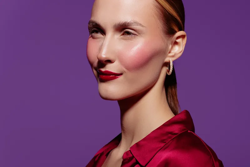 Portrait of confident woman with red lipstick and shiny skin wearing red blouse and gold hoop earrings on purple background. Concept of beauty, fashion, and elegance.