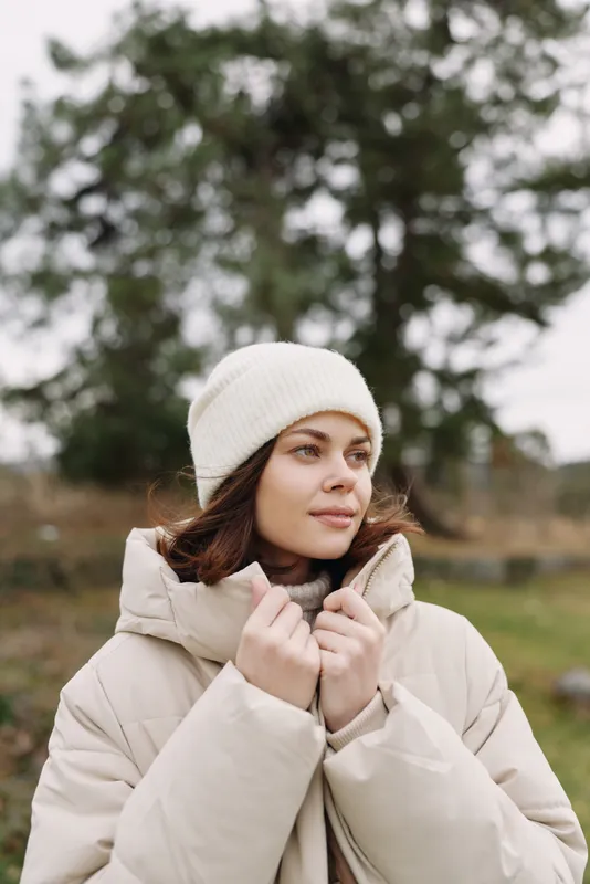 Woman in a warm beige coat and white beanie standing outdoors in a chilly countryside, calm expression, soft natural light and misty background, fashion and comfort in cold weather.