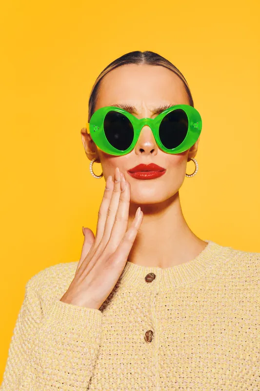 Stylish woman wearing large green sunglasses and red lipstick poses against bright yellow background. She wears a beige sweater and gold hoop earrings. Concept of fashion and summer style.