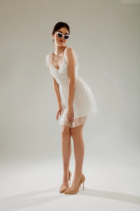 Elegant woman in a white lace dress and high heels poses confidently wearing stylish sunglasses. Studio shot on a neutral background. Concept of woman beauty fashion and elegance.
