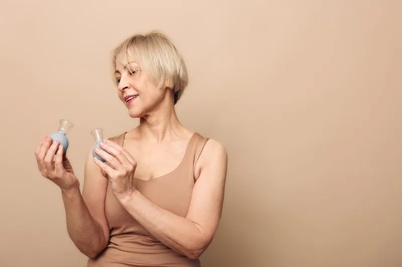 Smiling mature woman with short blonde hair wearing beige tank top holds two cosmetic bottles, examining skincare products on neutral background. Concept of beauty care and aging skin.