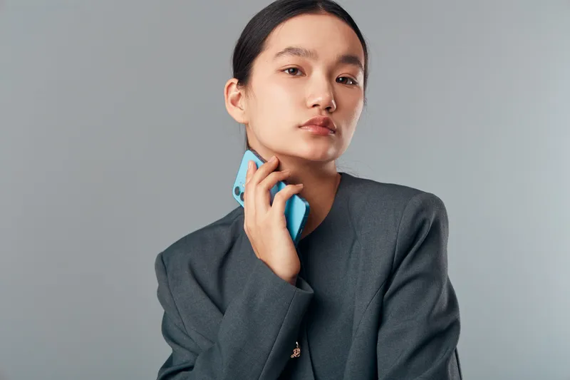 Portrait of confident young woman holding smartphone near face with hand, wearing dark blazer, posing against neutral grey background for technology, communication and fashion concept.