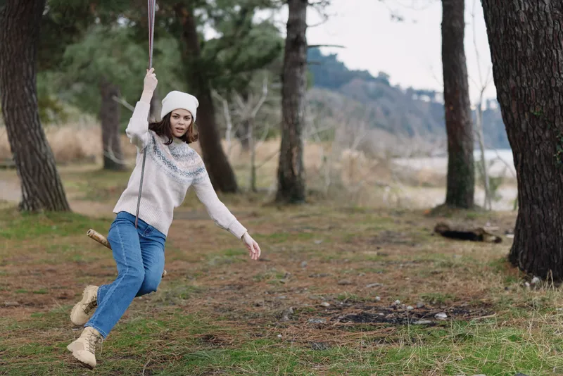 Outdoor girl on a swing in a forest, twirling with joy, wearing a cozy sweater, beanie and boots, with trees and grass around, a playful moment in nature and outdoor recreation