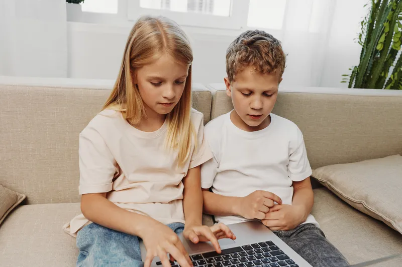 Two focused children sitting on sofa using laptop together in bright living room, learning and exploring digital technology concept for education and teamwork.