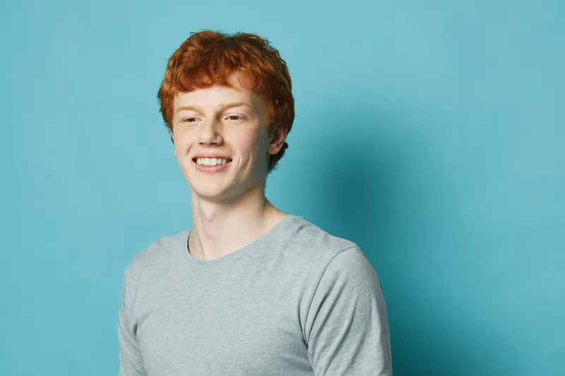 Young man with red hair and freckles smiling wearing gray shirt against blue background studio portrait of happy male model.