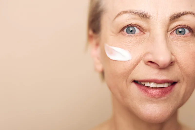 Smiling mature woman with blue eyes applies white face cream on cheek close up on beige background. Concept of skincare, anti aging, beauty, self care, and healthy skin routine.