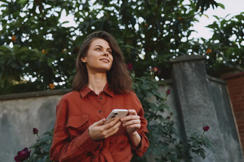Woman in red shirt stands outdoors holding a smartphone, capturing a casual lifestyle moment in a garden, looking up with a serene and confident expression