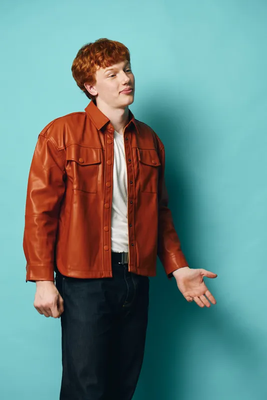 Young man with red hair wearing brown leather jacket and white shirt stands posing on blue background, casual style with thoughtful expression and relaxed posture.