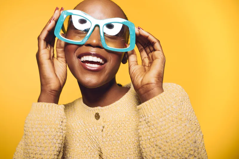 Happy black woman with shaved head wearing oversized blue glasses and beige sweater smiles brightly on yellow background. Concept of joy, fashion, and confident African ethnicity.