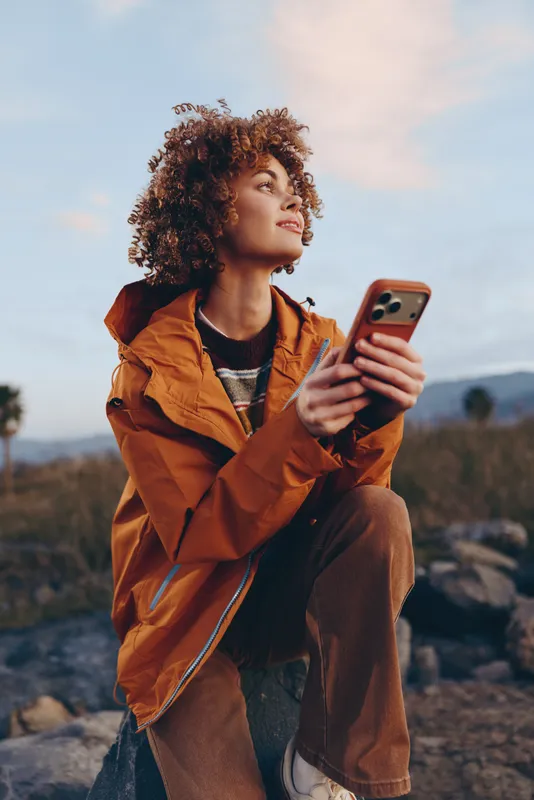 Woman with curly hair outdoors, wearing orange jacket, holding smartphone and looking up, lifestyle portrait in nature landscape, cloudy sky, soft natural light, relaxed mood, calmly