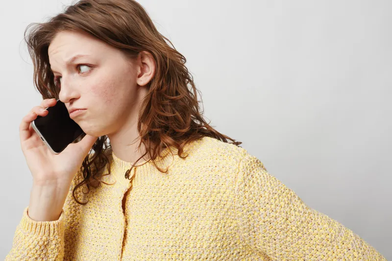 Young woman in yellow sweater looks worried while talking on phone against plain background. Concept of concern, communication, and emotional stress in everyday life.