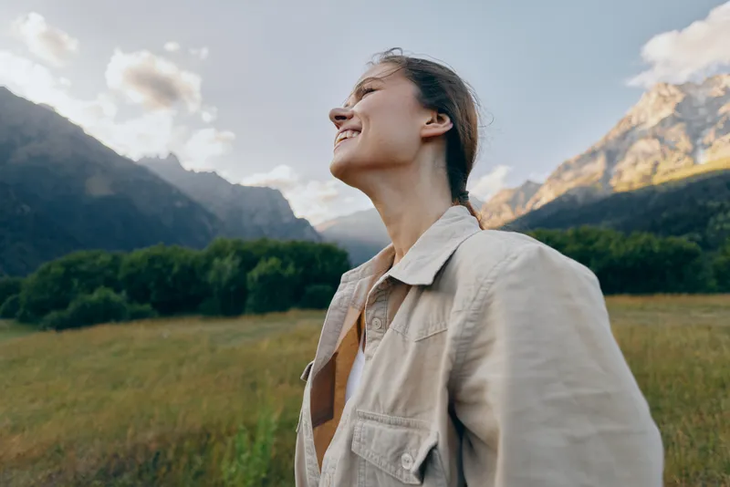 A smiling woman in a casual jacket stands in a field with distant mountains, enjoying daylight and nature, conveying happiness and a carefree outdoor lifestyle.