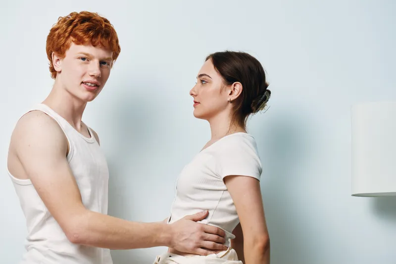 Young man and woman in casual white clothing standing close together in bright room with simple background, facing each other with relaxed expressions and gentle touch.