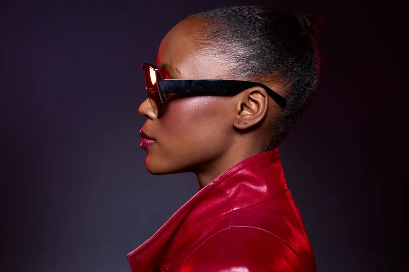 Profile of a stylish dark skinned African woman wearing large sunglasses and a red leather jacket against a dark purple background. Concept of fashion, elegance, and modern style.