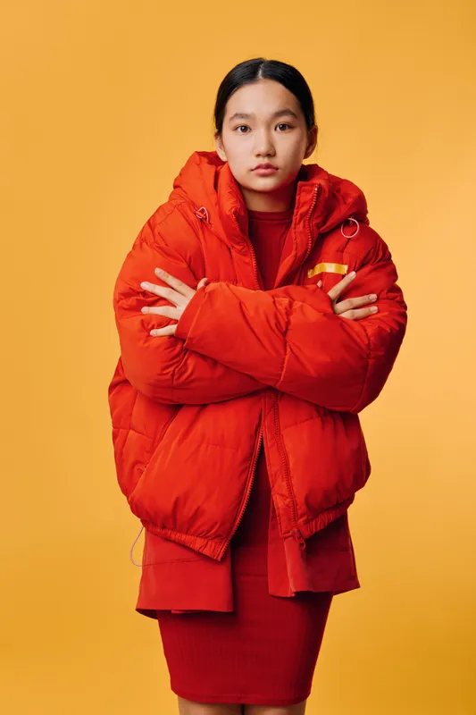 Young woman standing with crossed arms wearing red puffer jacket and matching dress against yellow background. Confident pose with casual fashion and winter outfit in studio.