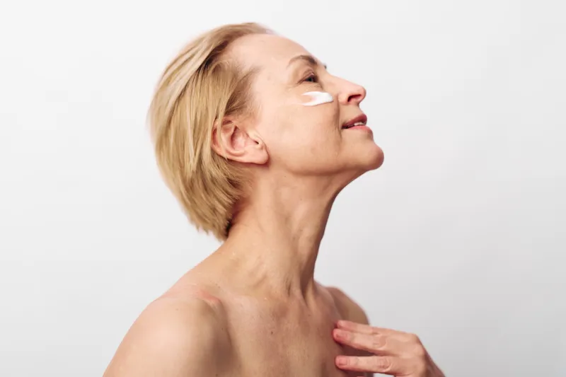 Mature woman with short blonde hair applies white cream under her eyes, tilting head back with eyes closed. Concept for skincare, antiaging, beauty, and selfcare routine.