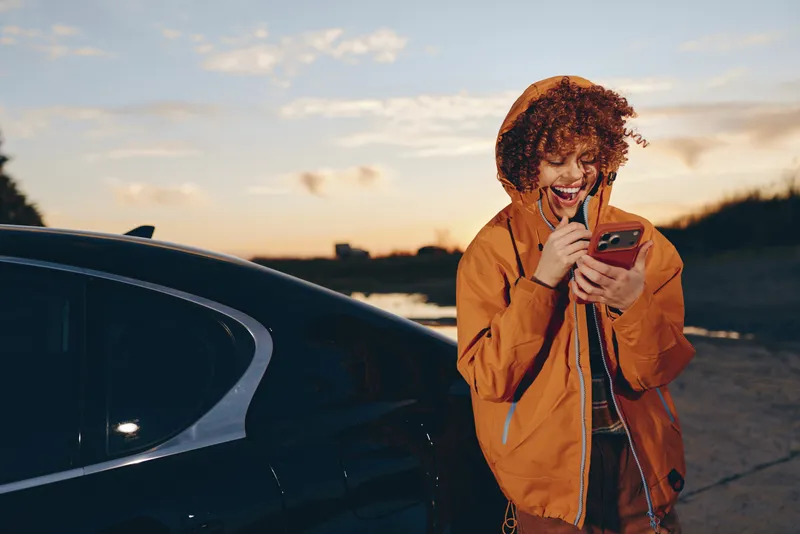 woman lifestyle outdoors at sunset near a car, smiling while looking at a phone, casual jacket outfit, candid travel mood, golden sky and open road evening breeze, relaxed stance