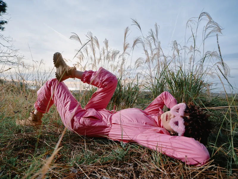 Outdoor fashion in a sunny meadow features a woman in a pink jumpsuit lying among tall grasses, relaxed pose, playful mood, and natural light emphasizing texture and color.