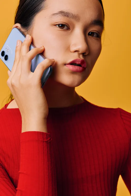 Young woman holding smartphone close to ear, talking or listening with focused expression, wearing red sweater against yellow background in studio portrait closeup.