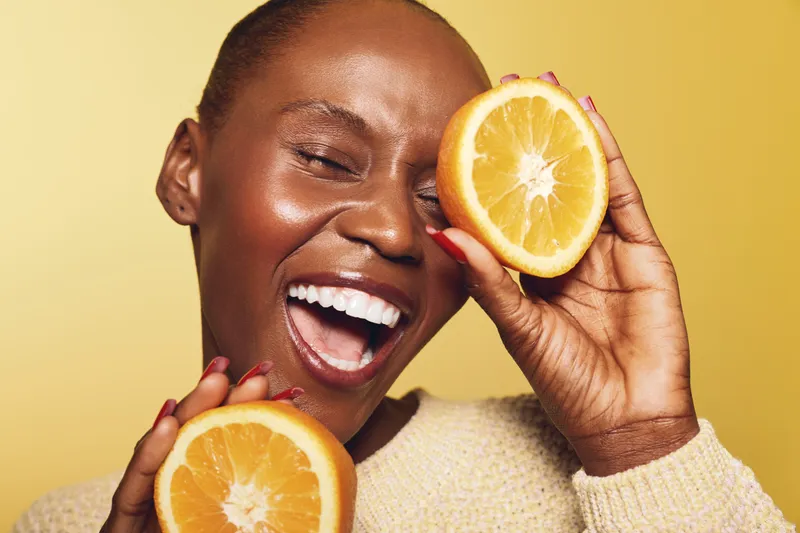 Happy African woman with dark skin holds orange halves near face laughing with closed eyes on yellow background. Concept of joy, healthy lifestyle, fruit, and natural beauty close-up.