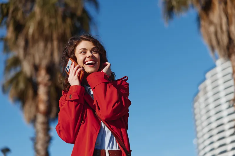 Smiling young woman wearing a red jacket talks on her smartphone outdoors under a clear blue sky, with palm trees behind her and a bright urban vibe