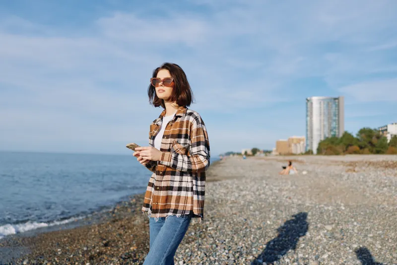 woman, beach, smartphone, casual, Outdoor scene of a woman using a smartphone on a sunny coastal beach, fashion plaid shirt and sunglasses with sea backdrop and relaxed mood.
