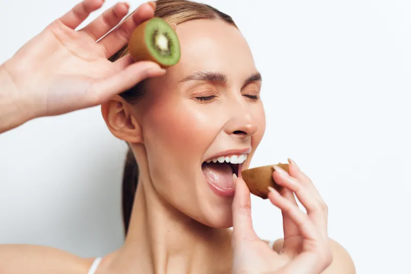 A young brunette woman tastes fresh kiwi, eyes closed in a playful moment, holding sliced kiwi near her mouth against a clean white background with a bright healthy glow