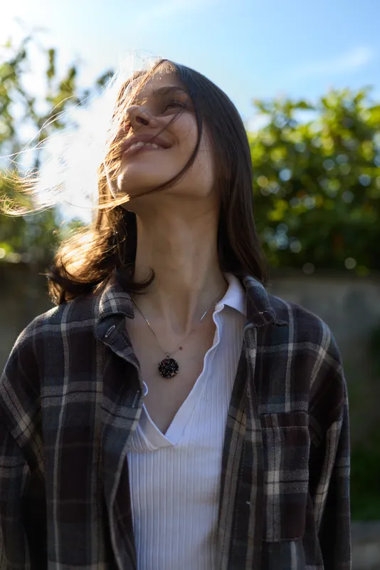 Candid woman outdoors, smiling with head tilted, sunlit moment, relaxed posture, breeze through hair, natural colors, lush greenery background, plaid shirt over white top, casual lifestyle vibe