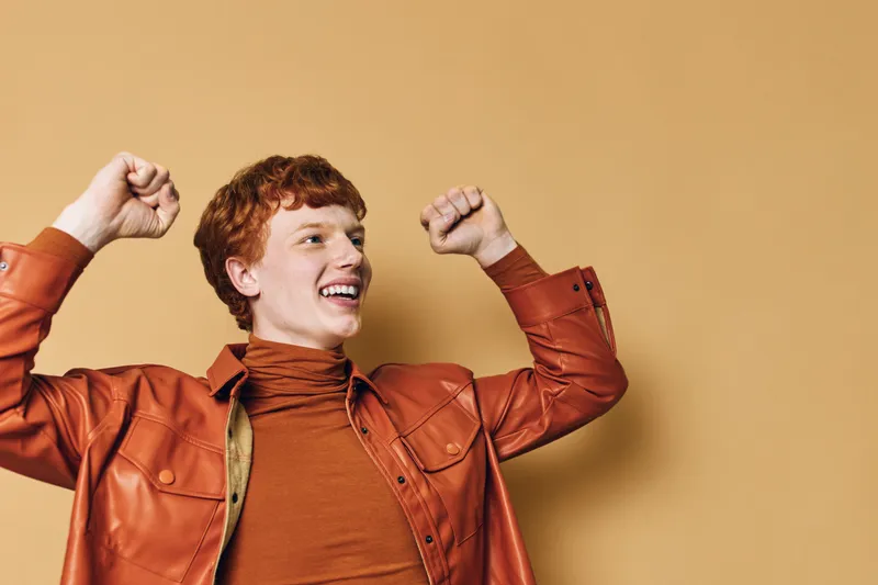 Young redhead man wearing leather jacket celebrating with raised fists and smiling isolated on light brown background for success and joy expression.