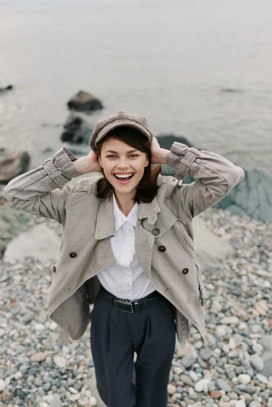 Happy young woman in casual jacket and hat smiles broadly, standing on rocky beach near water. Outdoor lifestyle, fashion, and joyful expression concept in natural light.