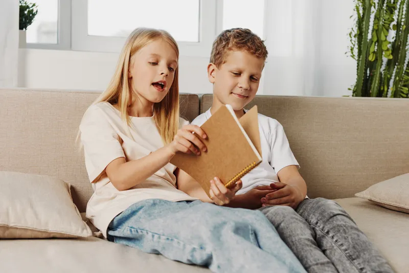 Two children sitting on a couch reading a notebook together in a bright living room. Concept of learning, childhood, and shared curiosity.