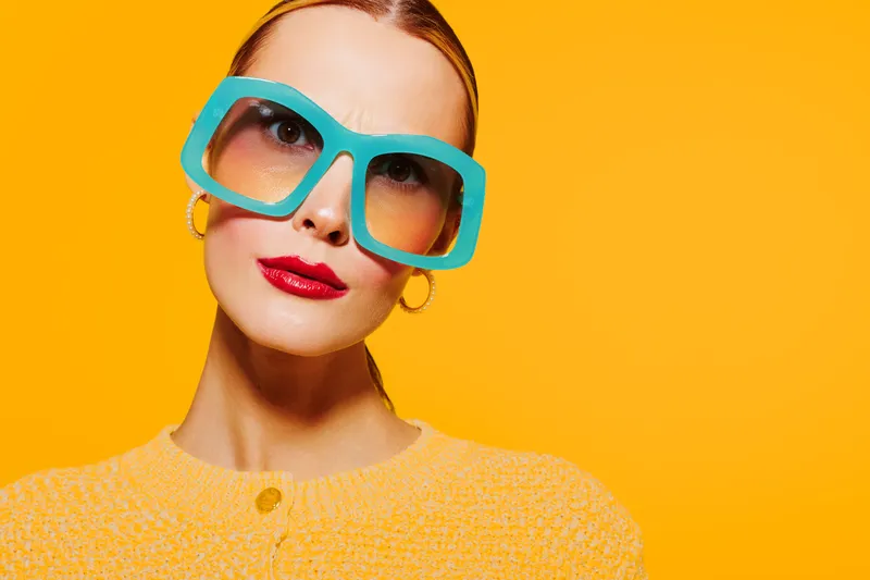 Stylish woman with large turquoise sunglasses and red lipstick poses against bright orange background wearing yellow sweater. Concept of fashion, beauty, and vibrant colors.