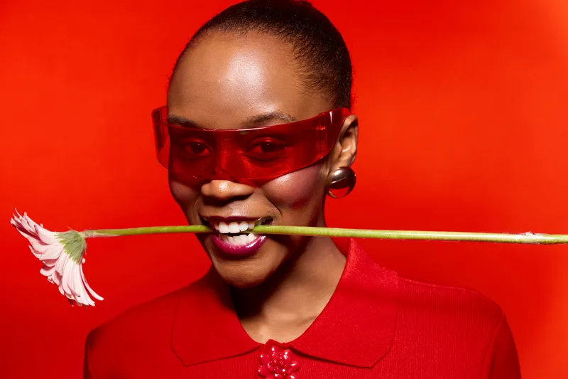 Smiling African woman with dark skin wearing red sunglasses and red outfit holds pink flower in mouth against bright red background. Concept of fashion, joy, and vibrant color contrast.