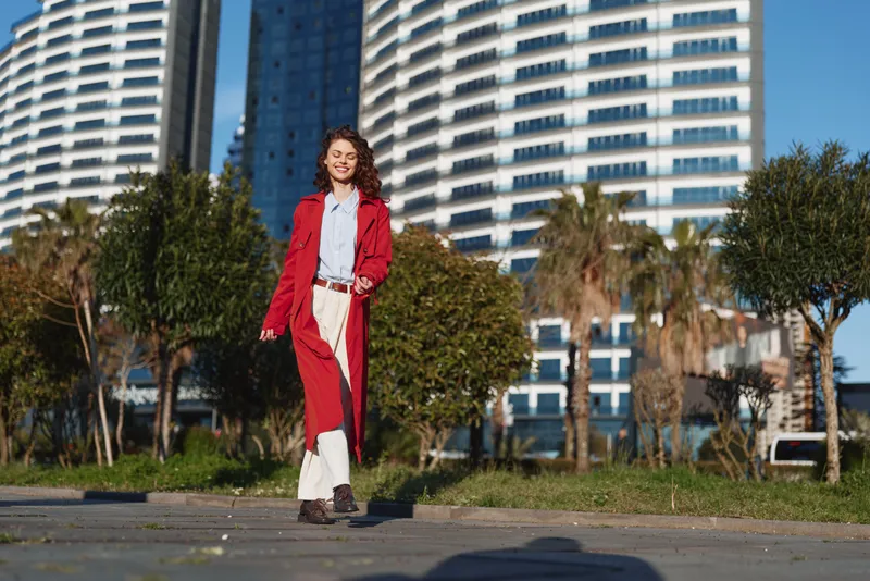 Young woman walking outdoors in a red coat and white pants, smiling with eyes closed, urban background with modern buildings and palm trees, sunny day, casual style.