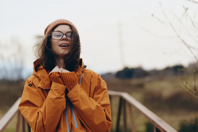 Cheerful woman outdoors, laughing in winter sunlight, wearing orange jacket, knit hat, scarf and glasses, relaxed lifestyle moment on a countryside path, warm colors, soft breeze, blurred nature