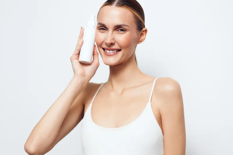 Smiling woman holding white cosmetic bottle near face, clear skin, natural makeup, simple white top, bright studio background, skincare concept, beauty routine, fresh appearance